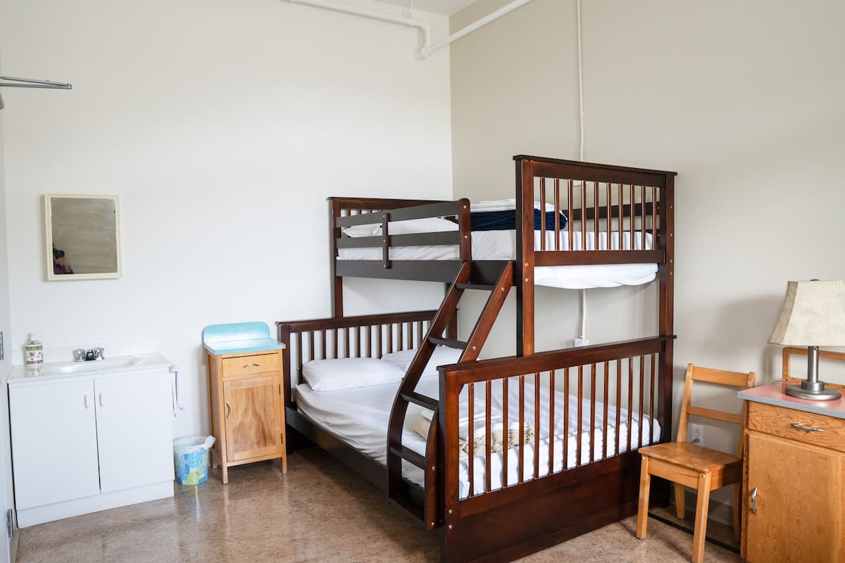 Habitación con literas, cabecera de madera, una silla pequeña de madera, escritorio con lámpara, armario blanco y un basurero.