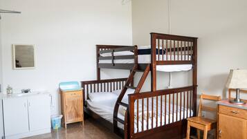 Habitación con literas, cabecera de madera, una silla pequeña de madera, escritorio con lámpara, armario blanco y un basurero.