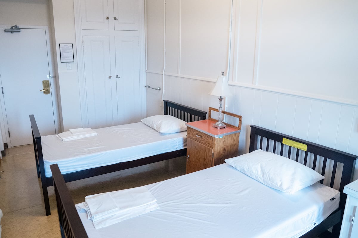 Una habitación con dos camas individuales, una mesita de noche de madera con lámpara y una puerta blanca con manija.