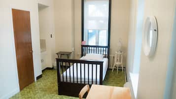 A small room with a single bed, a wooden table, a chair, a window with blinds, a radiator, and a door.
