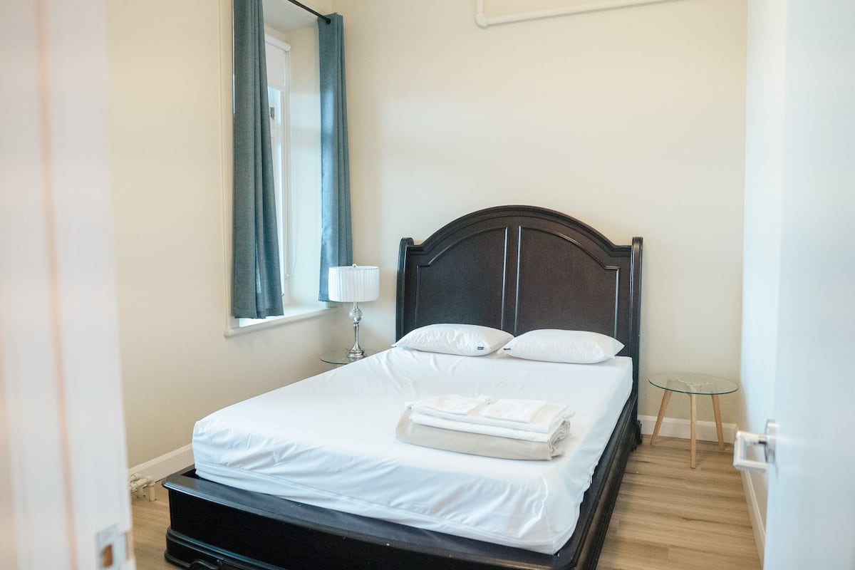 Una cama bien hecha con sábanas blancas y una toalla doblada, una mesita de noche con una lámpara y una pequeña mesa de vidrio junto a la cama.