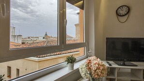 Une pièce avec une grande fenêtre, un bureau avec un ordinateur, une horloge au mur et une plante en pot sur le rebord de la fenêtre.