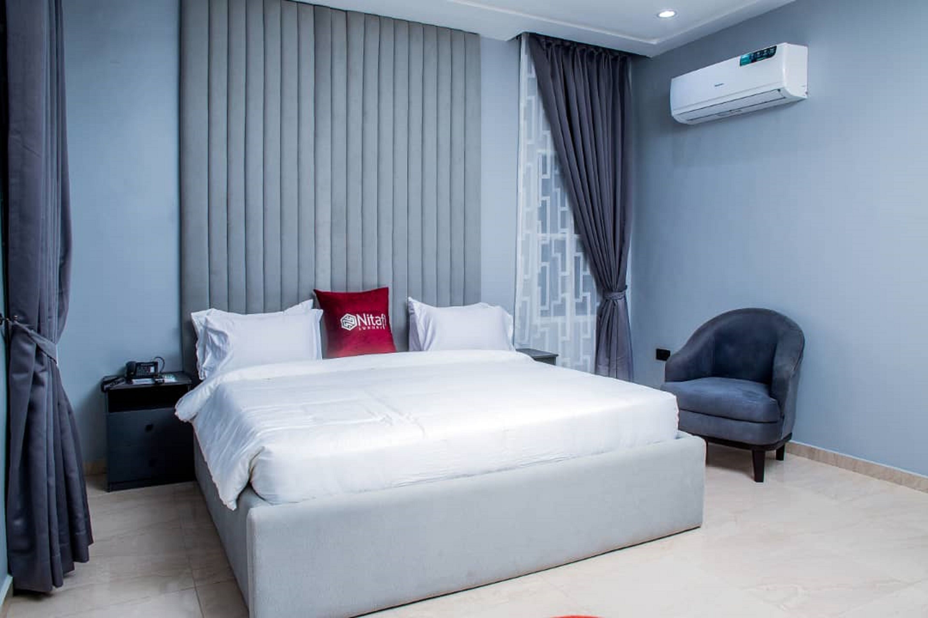 A modern bedroom with a large bed, grey curtains, a grey armchair, and an air conditioning unit on the wall.