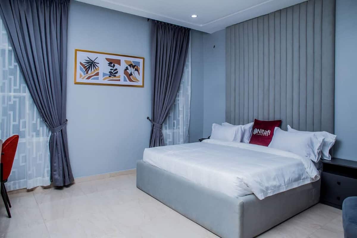 Un dormitorio con cama, cortinas grises, almohada roja y un cuadro enmarcado en la pared.