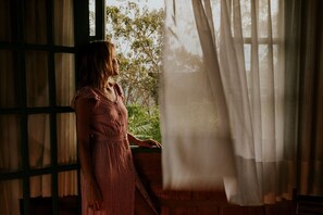 Una persona con vestido rojo de pie junto a una ventana con cortinas traslúcidas, con vista a un jardín.