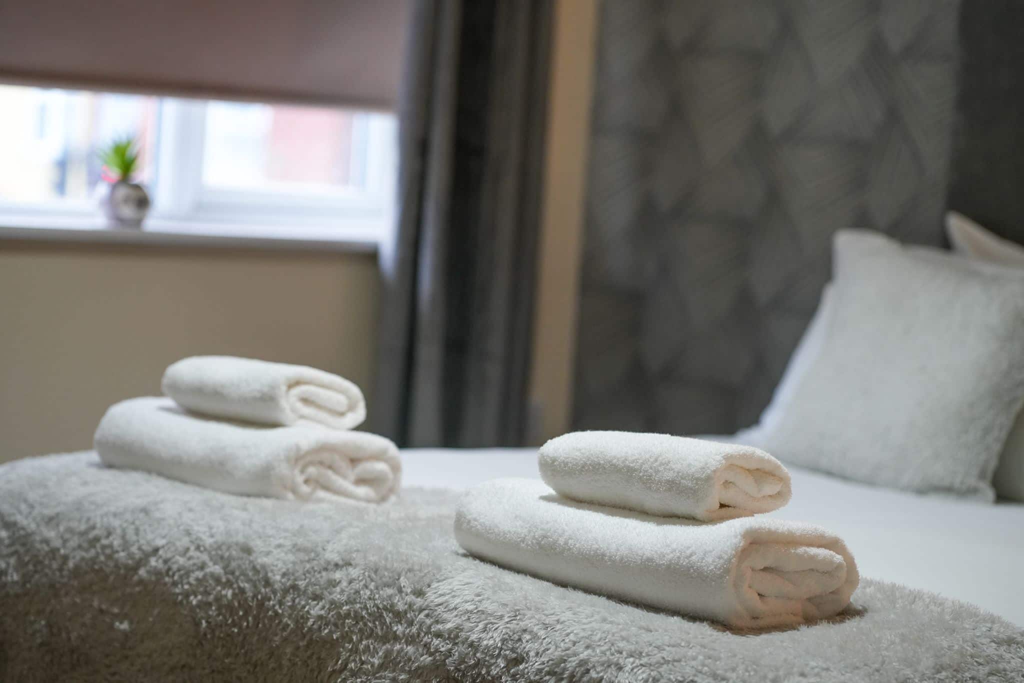 Un lit bien fait, avec des serviettes blanches pliées en rouleaux et une couverture grise à texture intéressante.
