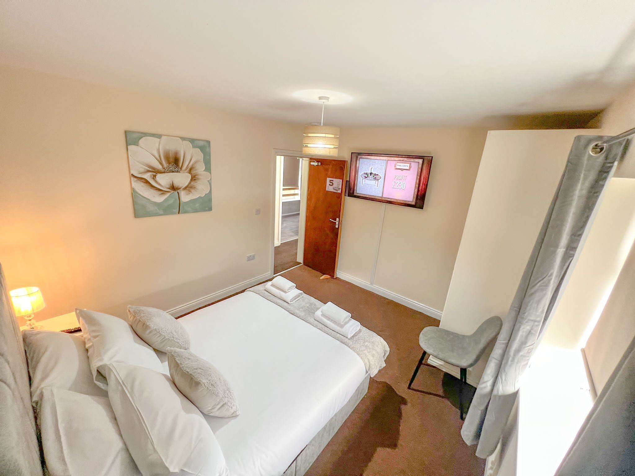 A hotel room with a large bed, a desk, a chair, a wall-mounted TV, and a door with a number sign.