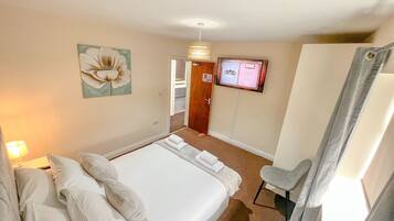 A hotel room with a large bed, a desk, a chair, a wall-mounted TV, and a door with a number sign.