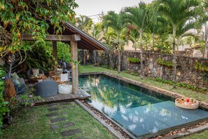 A swimming pool with a wooden deck, stone wall, and tropical plants.
