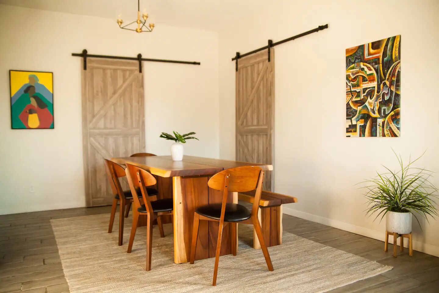 A dining area with a wooden table and chairs, a potted plant, and two abstract paintings on the walls.