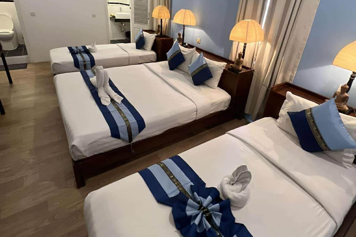 A hotel room with three beds, each with white linens and blue accents. There are lamps on each nightstand, and a ceiling fan is visible.