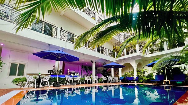 An indoor swimming pool with multiple levels, surrounded by white chairs and blue umbrellas, and adorned with palm trees. - Pride Villa Cambodia (Phnom Penh)