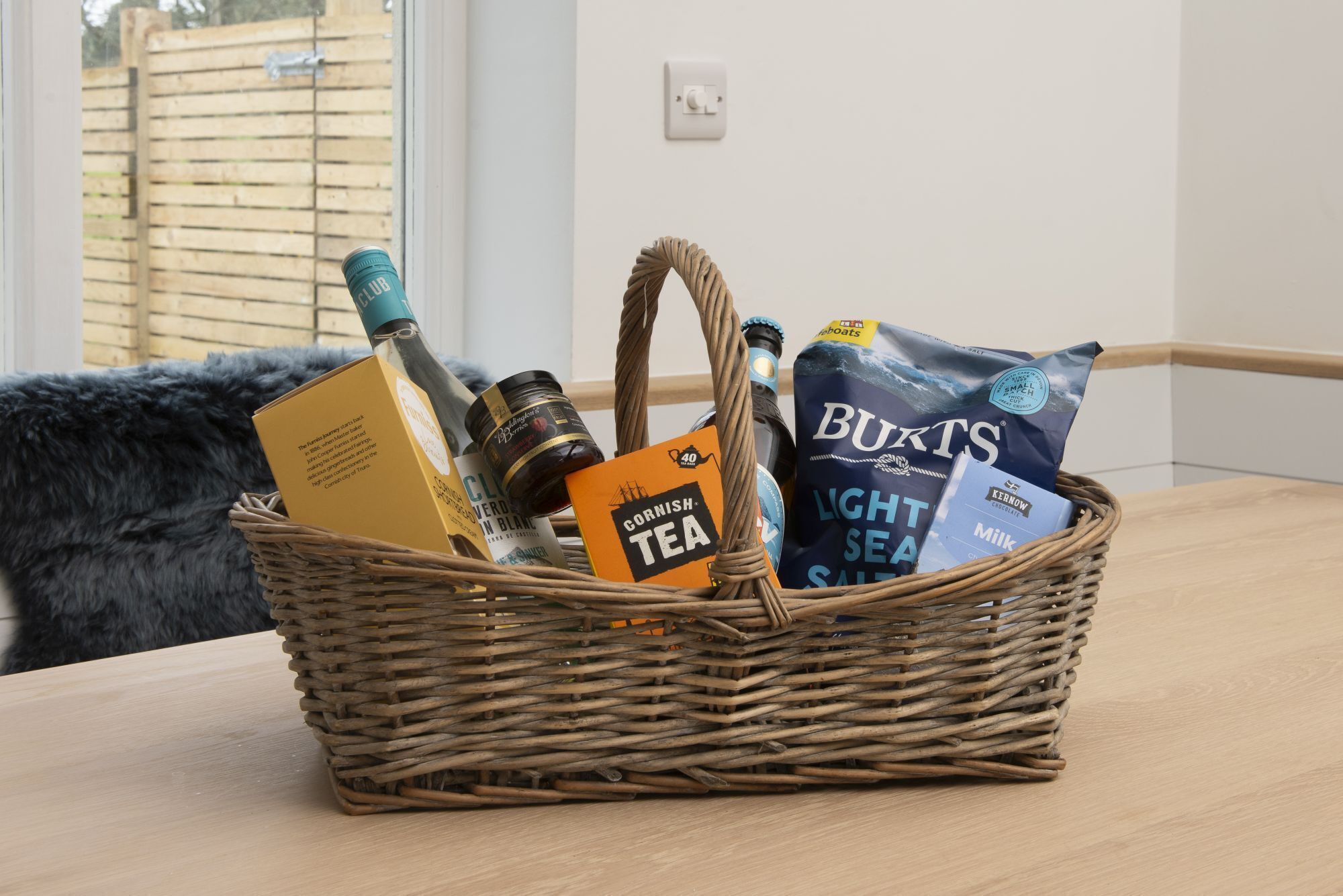 A wicker basket containing a bottle of beer, a box of tea, and a bag of milk on a wooden table.