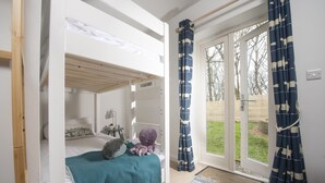 A bedroom with a bunk bed, a bedside table with a lamp, and a view of the garden through a sliding door. - Lobbs Cottage -  Pet Friendly, Free Parking (St Mawgan)