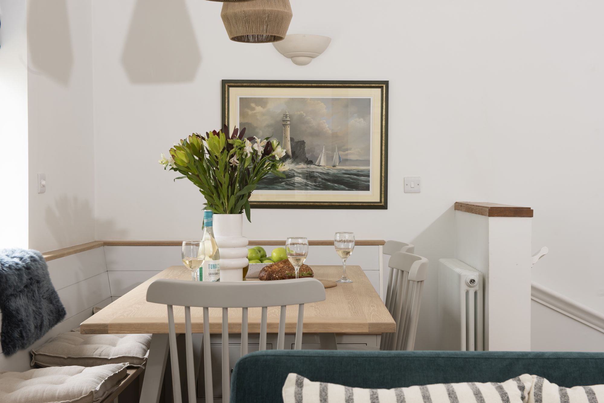 Een eetruimte met een tafel gedekt voor twee personen, een vaas met bloemen, een fles witte wijn en een lijst met een schilderij van een vuurtoren.