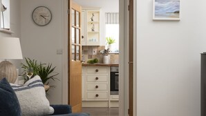 A modern living room with a sofa, a clock, and a view into a kitchen with wooden cabinets and a window. - Lobbs Cottage -  Pet Friendly, Free Parking (St Mawgan)