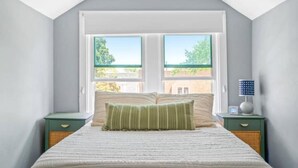 A bedroom with a large window, a bed with striped bedding, two bedside tables, and a lamp on one table. - The Norwood Nook – Walkable Stay Near O’Hare & Metra! (Chicago)