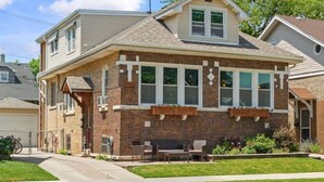 A two-story house with a brick facade, large windows, and a front porch with outdoor seating. - The Norwood Nook – Walkable Stay Near O’Hare & Metra! (Chicago)