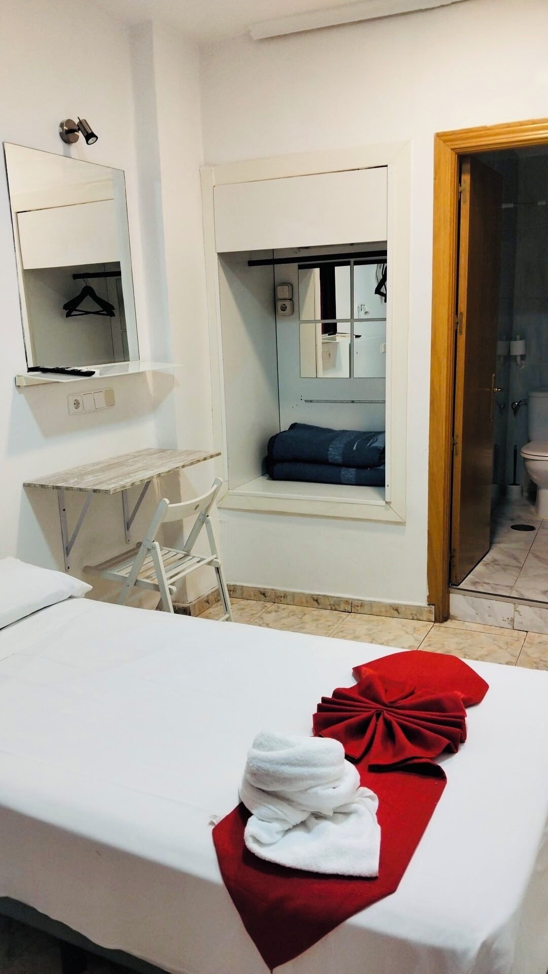 A hotel room with a bed, a desk, a chair, a mirror, a window, and a bathroom visible through the door.