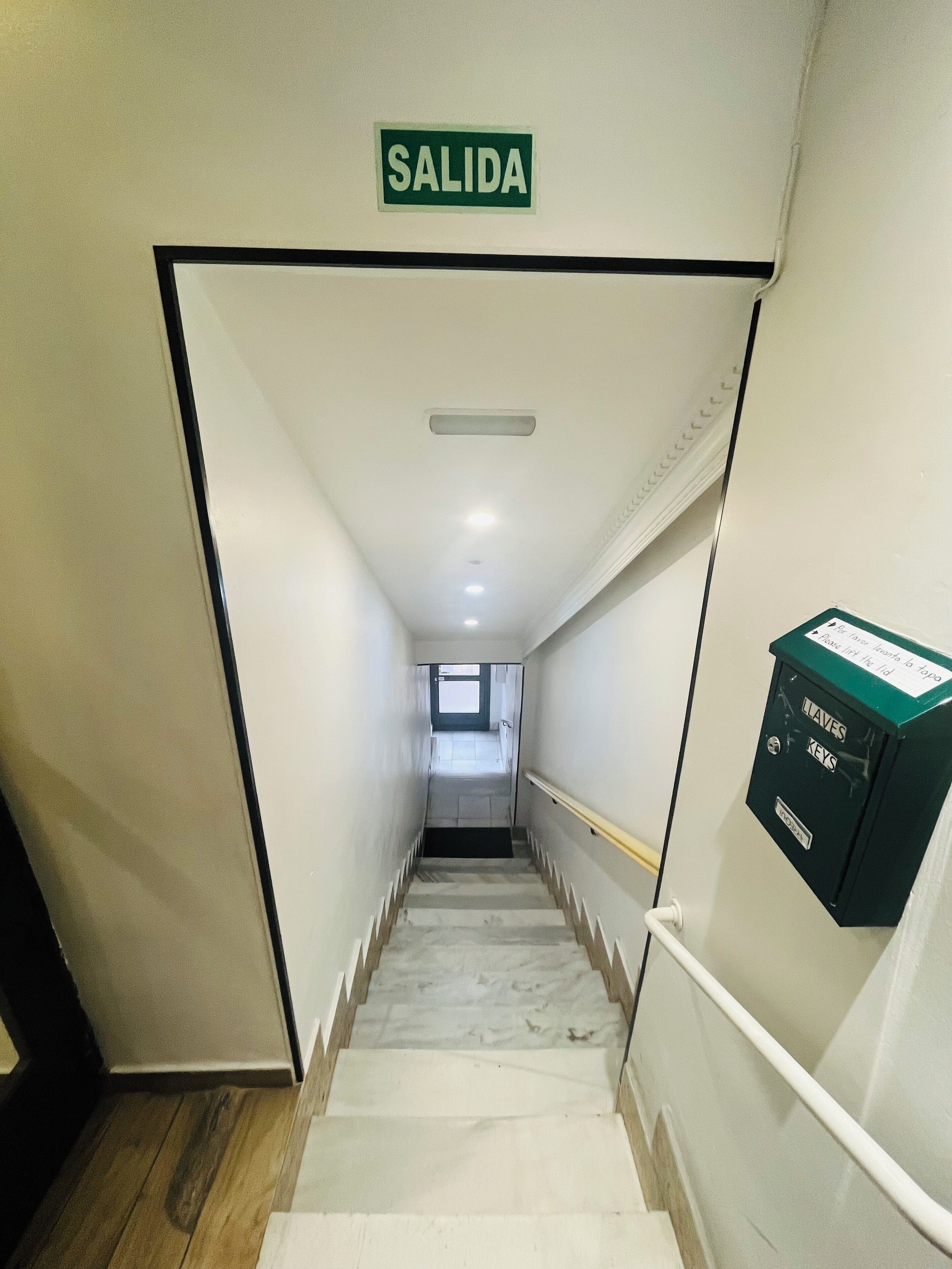 A hallway with a sign reading "SALIDA" at the top, a staircase with a handrail, and a box labeled "LLAVES KEYS" on the right wall.