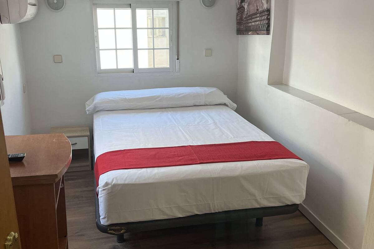 Un dormitorio con cama, un escritorio de madera, una mesita de noche, una ventana y un aire acondicionado instalado en la pared.