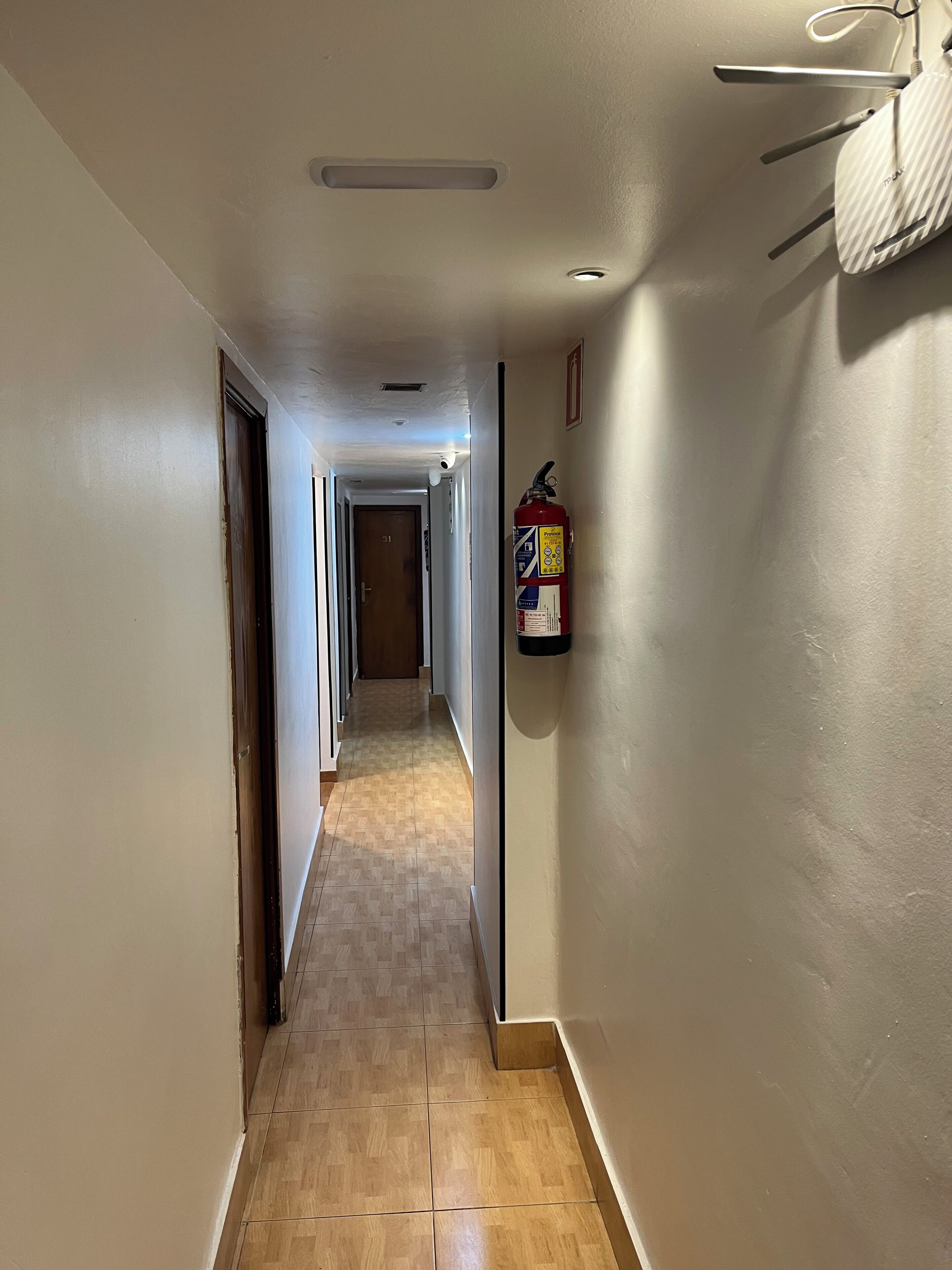 A hotel hallway with a fire extinguisher mounted on the wall, a ceiling light, and a door at the end.