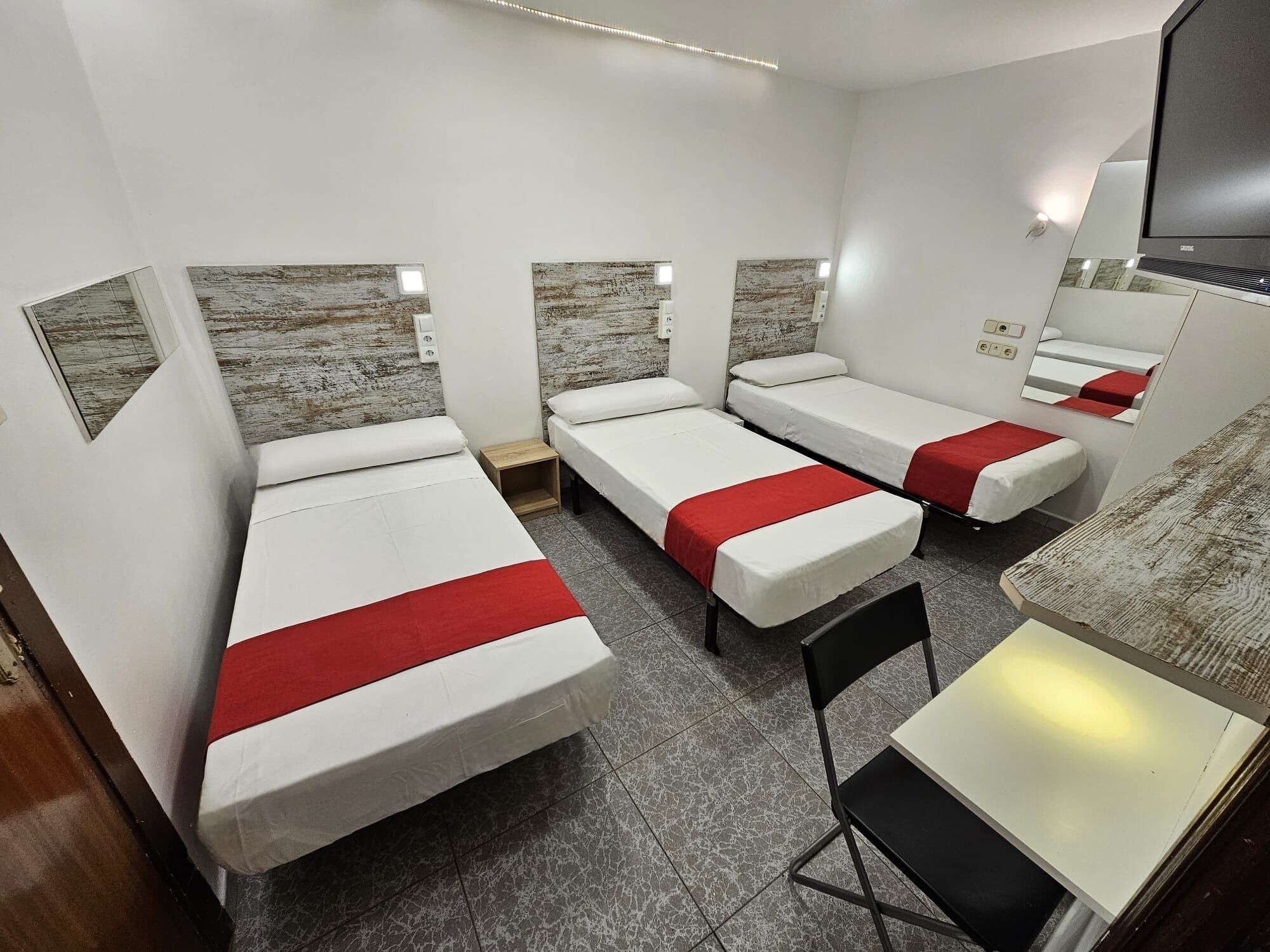 A hotel room with three single beds, a small desk, a chair, a television, and a mirror.