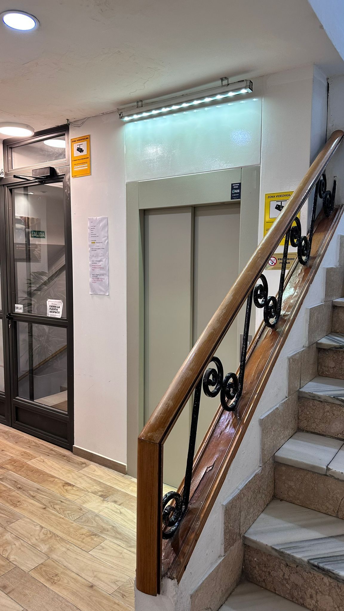 A staircase with wooden handrails and a metal banister, leading to a landing with a glass elevator door and a closed elevator.