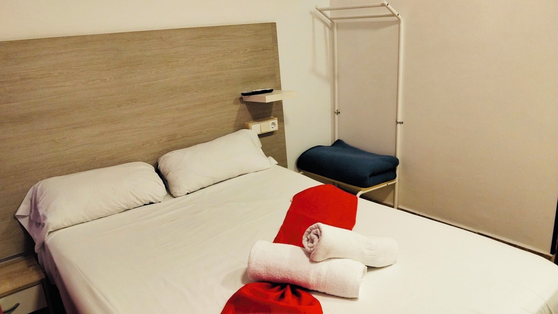 Un lit bien fait, avec du linge de lit blanc et deux oreillers, une serviette rouge et une chaise bleue, dans une chambre simple dotée d’une tête de lit en bois et d’une armoire blanche.