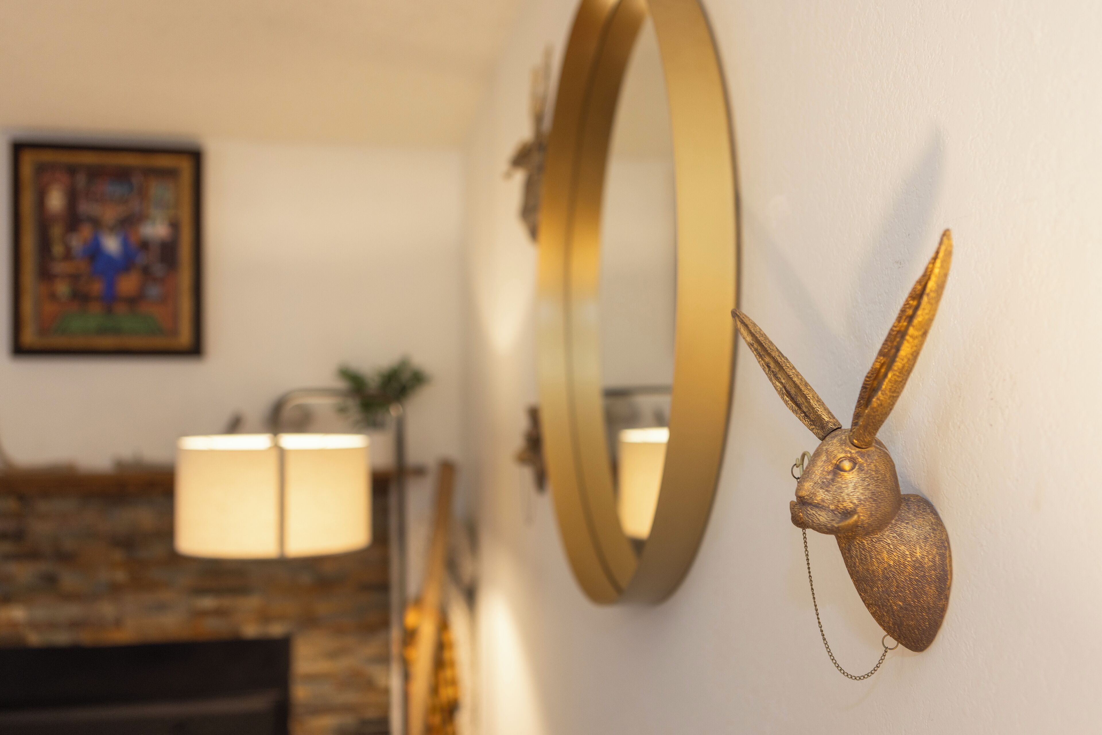 A wall-mounted mirror with a golden frame, a decorative rabbit head, and a framed picture in the background.