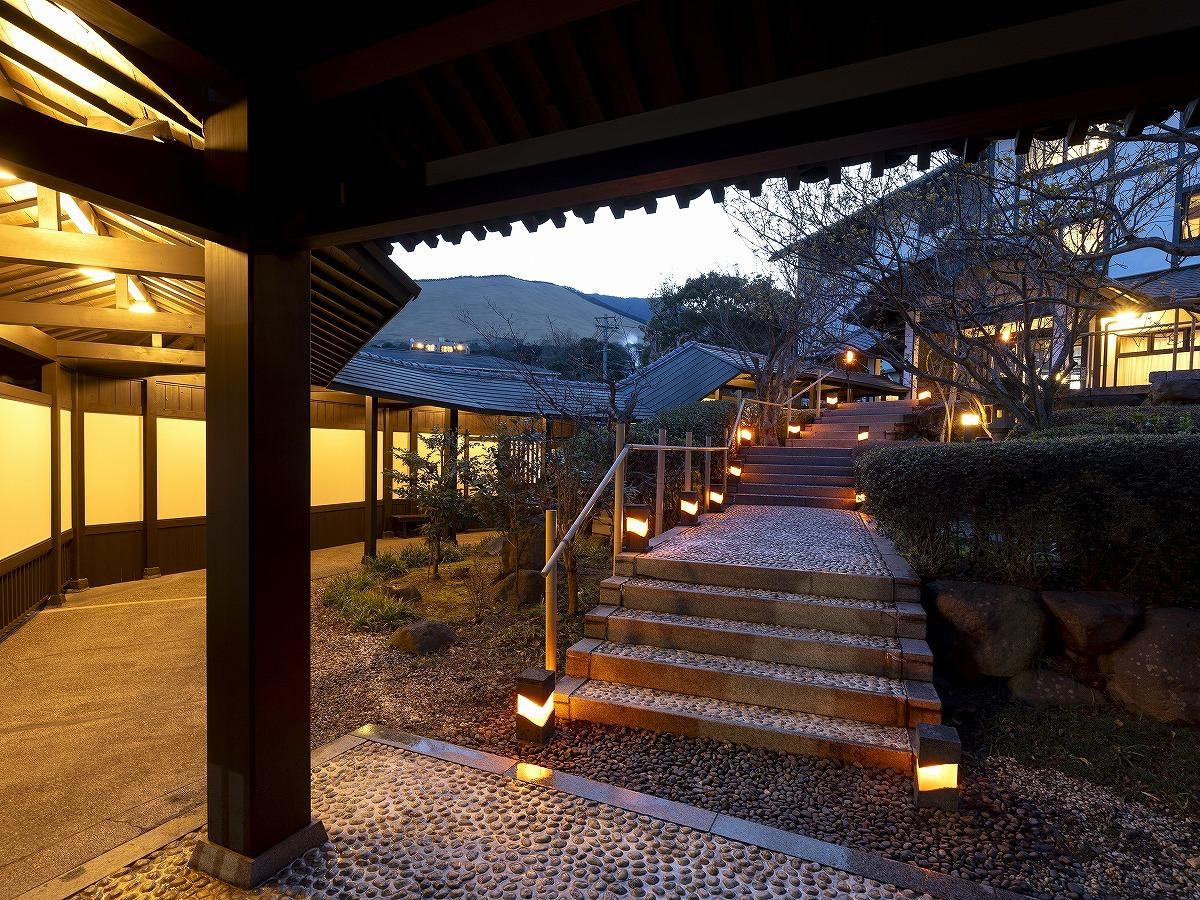Un edificio tradicional japonés con estructura de madera, un sendero de piedra que conduce a un conjunto de escalones y una vista de una montaña al fondo.