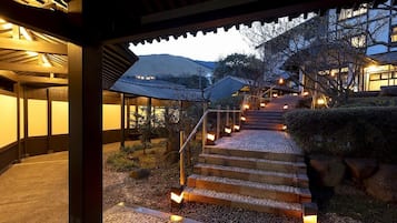 Un edificio tradicional japonés con estructura de madera, un sendero de piedra que conduce a un conjunto de escalones y una vista de una montaña al fondo.