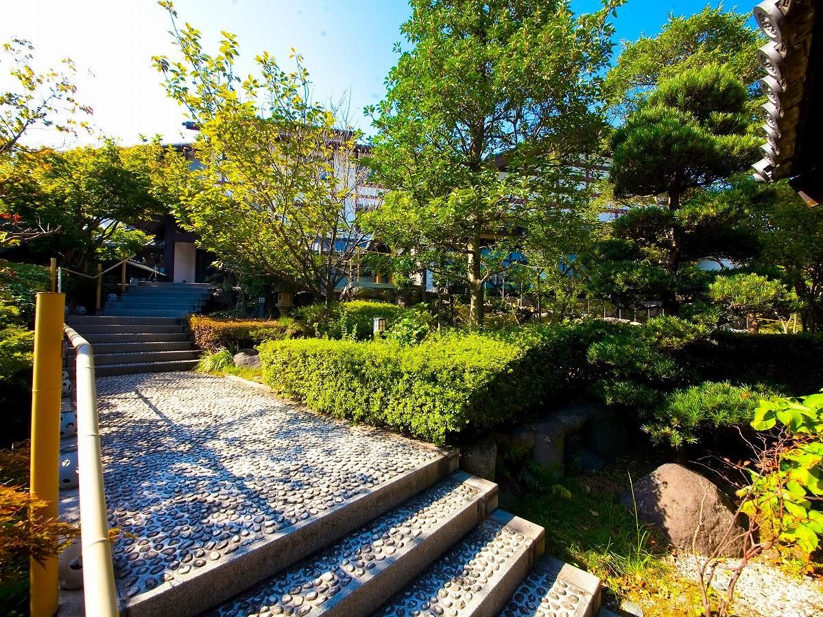 Un sendero de piedra conduce a un edificio de arquitectura tradicional, rodeado de abundante vegetación y un cielo despejado.
