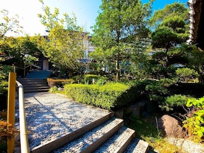 A stone pathway leading to a building with traditional architecture, surrounded by lush greenery and a clear sky.