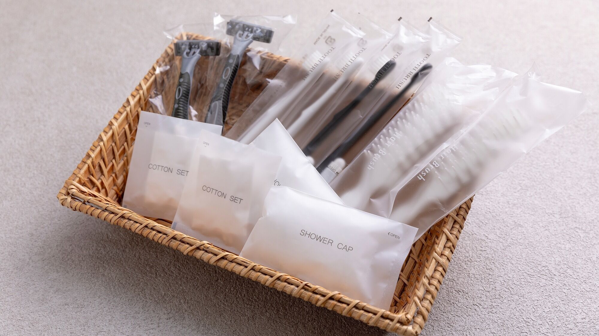 A wicker basket containing several plastic bags with items labeled 'COTTON SET' and 'SHOWER CAP.'