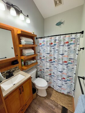 A bathroom with a wooden vanity, a white sink, a toilet, and a shower curtain with a fish pattern. - Cozy Getaway by the Lake
 (Twin Lakes)