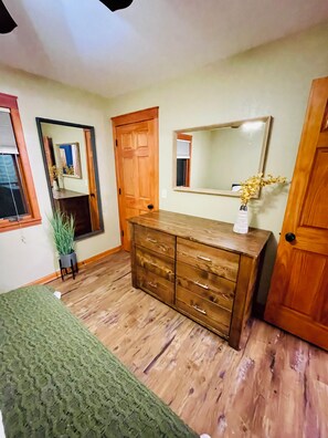 A room with wooden flooring, a green knitted area rug, a wooden dresser with drawers, a mirror above the dresser, and a potted plant. - Cozy Getaway by the Lake
 (Twin Lakes)