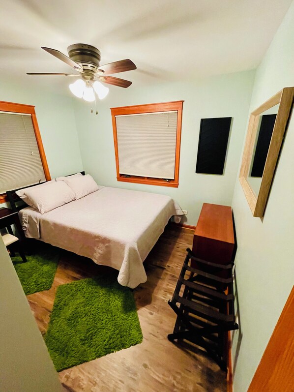 Ein kleines, ordentlich eingerichtetes Schlafzimmer mit Bett, Schreibtisch, Stuhl, Deckenventilator und einem Fenster mit Jalousien.