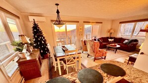 A spacious living area with a dining table, a Christmas tree, and a large window offering a view of the outdoors. - PanoView•Spa•PoolTable•Fireplace•2Kings•Pickleball•Arcade•Fire Table•Central (Seven Devils)