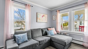 Un canapé d’angle gris avec des coussins bleus et roses, un tableau abstrait encadré et une fenêtre avec des rideaux roses.