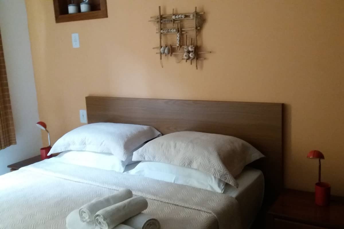 A neatly made bed with white linens and rolled towels, a wooden headboard, and a wall-mounted shelf with kitchen items.