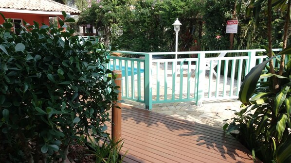 A wooden deck with a white fence, surrounded by lush greenery and a sign that reads "ATENÇÃO".