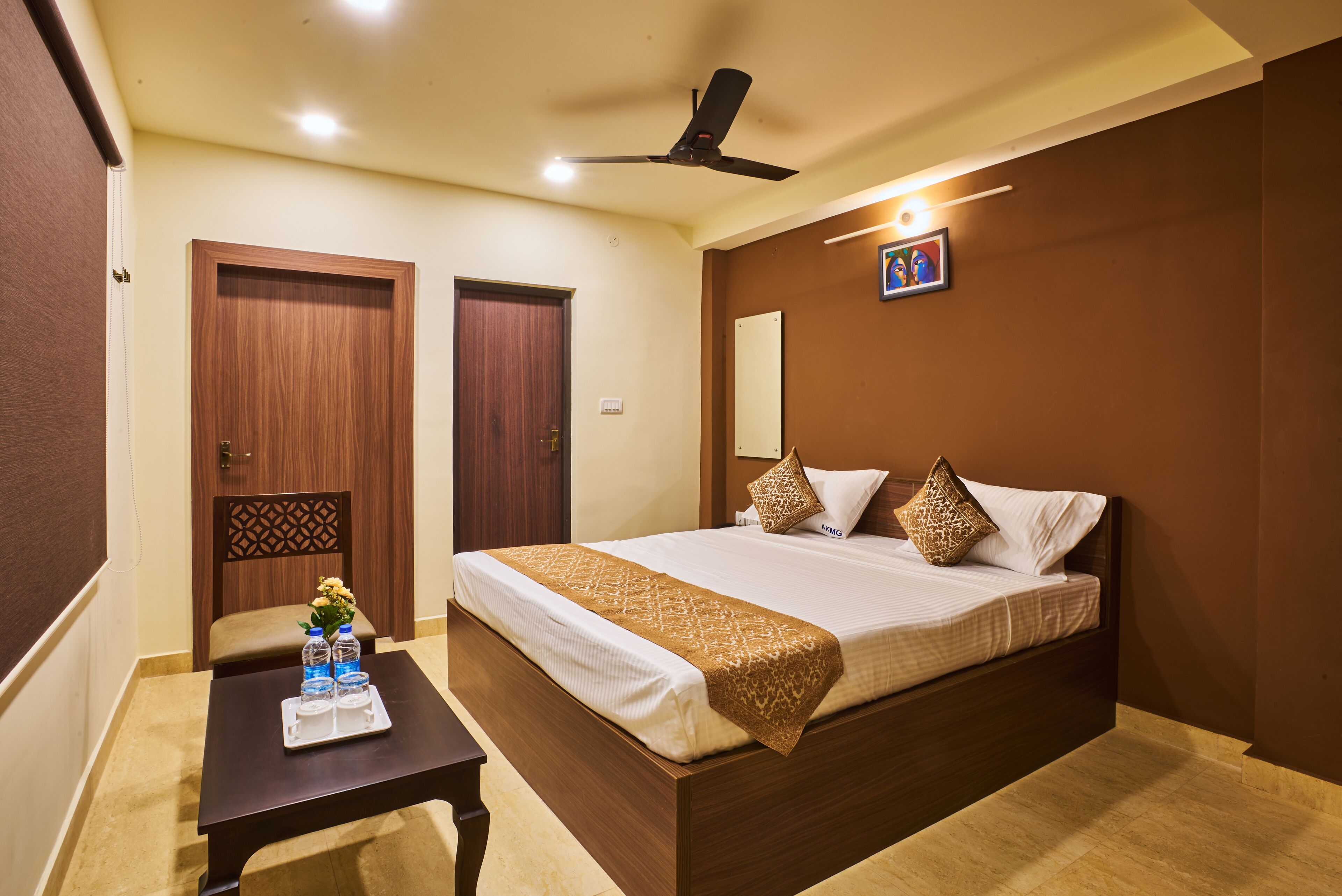 A hotel room with a large bed, a ceiling fan, a small table with water bottles, and a decorative pillow.