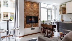 Ein modernes Wohnzimmer mit Ziegelwand, Flachbildfernseher, Essbereich und Küche mit dunklen Schränken.