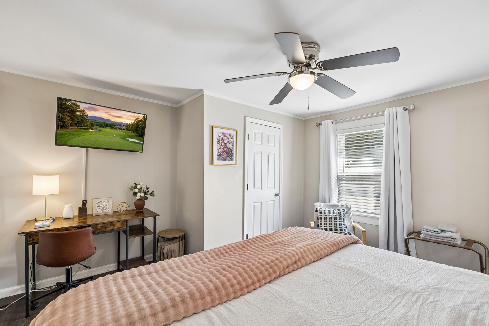 A hotel room with a bed, desk, chair, ceiling fan, TV, and a window with curtains.