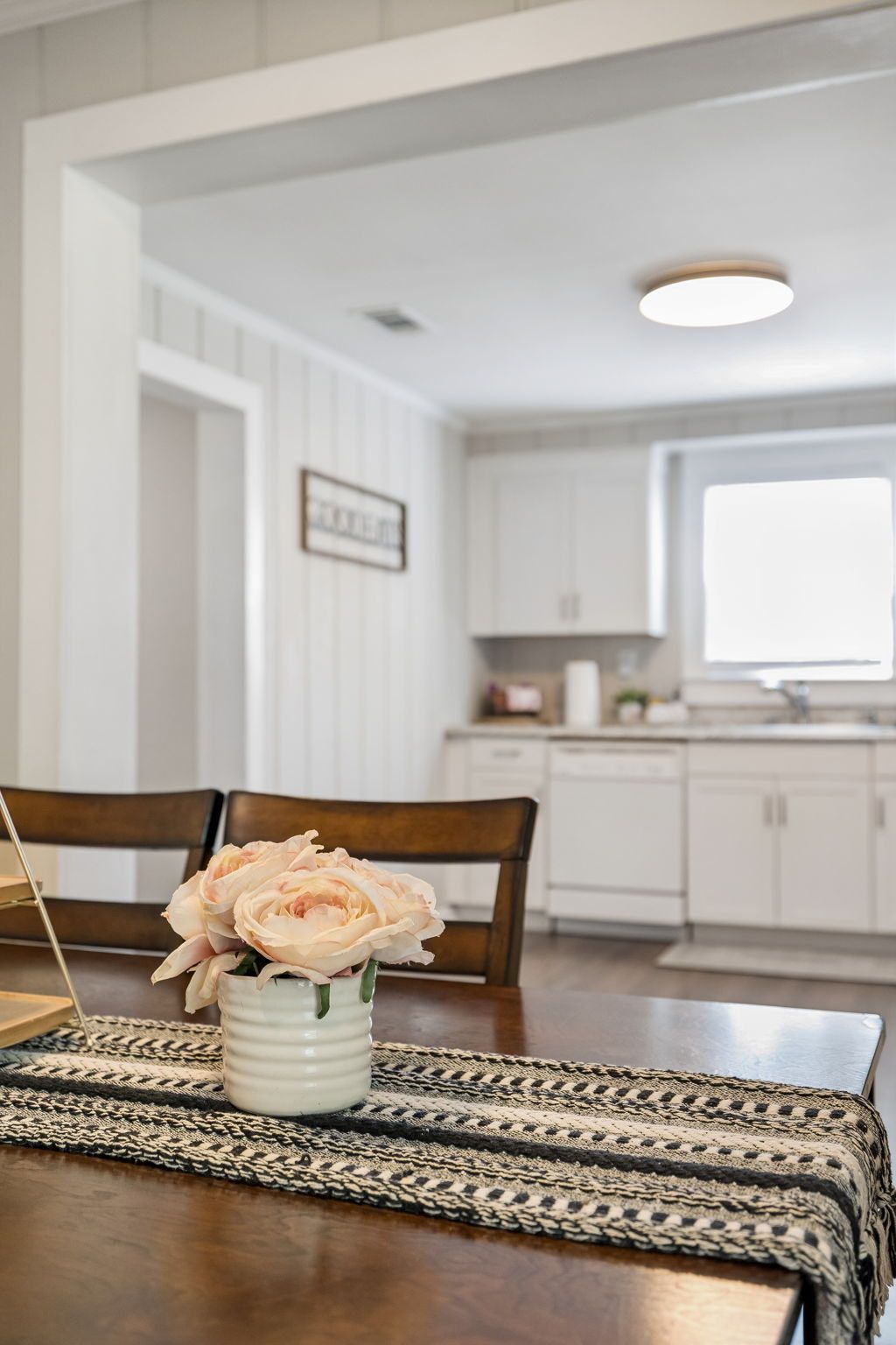 A dining area with a table set for two, a vase with flowers, and a kitchen in the background.