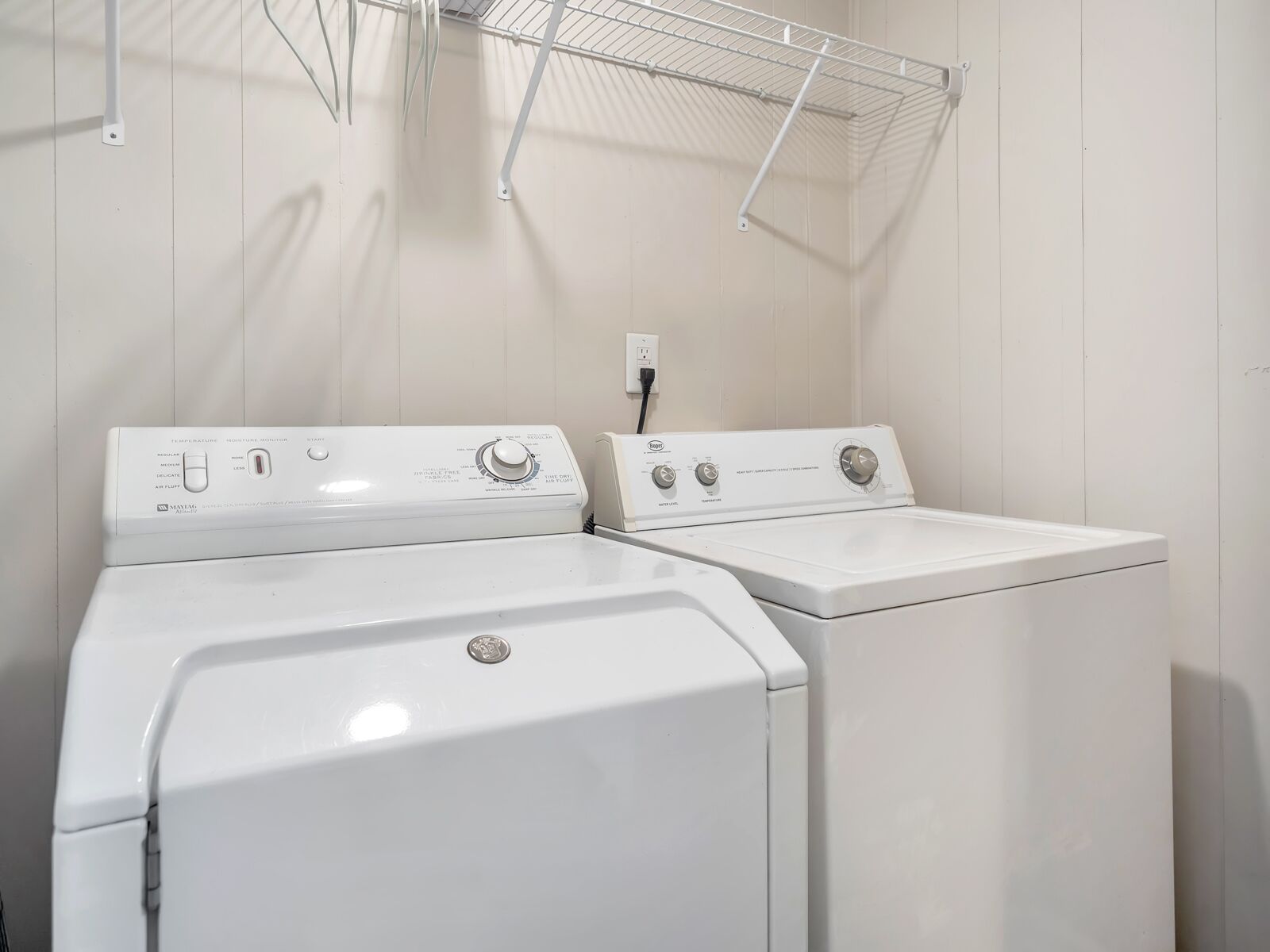 A laundry room with two white front-loading washers and dryers, a metal drying rack, and a wall-mounted electrical outlet.