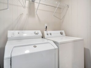A laundry room with two white front-loading washers and dryers, a metal drying rack, and a wall-mounted electrical outlet.