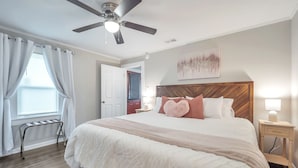 A bedroom with a large bed, a ceiling fan, a nightstand with a lamp, and a window with curtains. - Cute Modern Townhouse Close to MTSU, Parks & Downtown: Large Master & Coffee Bar (Murfreesboro)