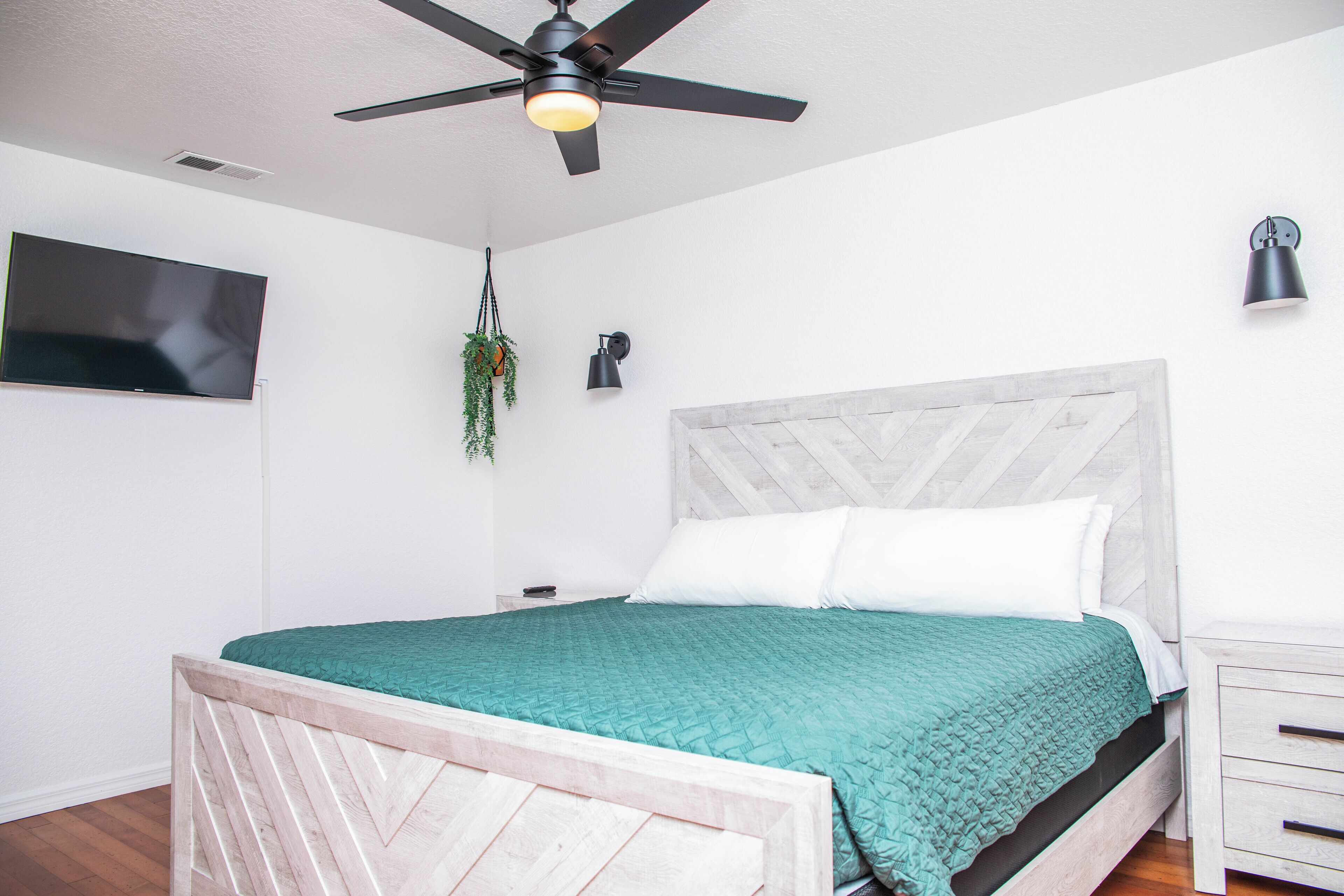 A bedroom with a bed, a ceiling fan, a TV mounted on the wall, and a bedside table with a lamp.